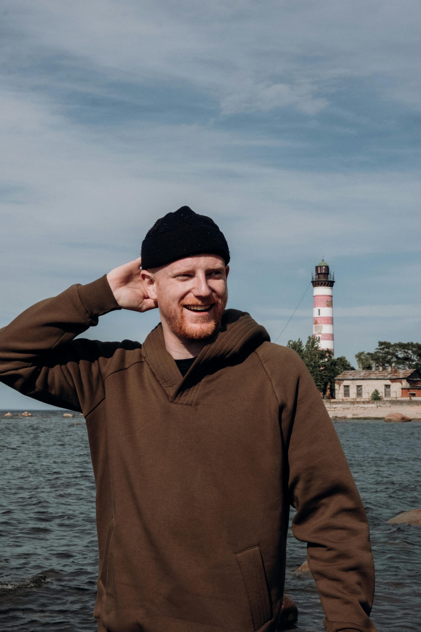 A cheerful man in a hoodie and beanie poses by the sea with a lighthouse in the background.